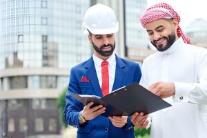 Two Arabic men in business suits and hard hats, discussing a project, representing a Kuwait job vacancy opportunity.