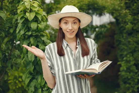 A woman in a hat holds an open book titled Environmental Literacy, engaged in reading outdoors.